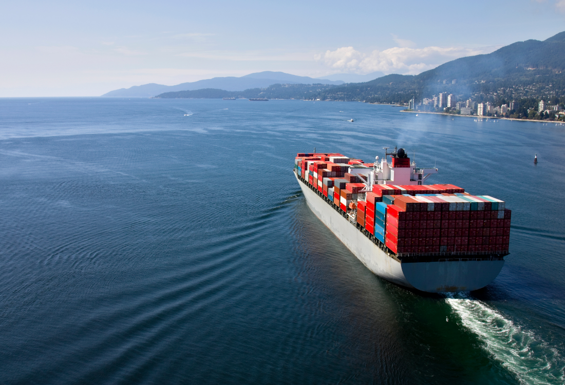 A large container ship sails through calm blue waters with mountains and a coastal city in the background, representing global shipping routes and the challenges facing Western vessels in the Red Sea.