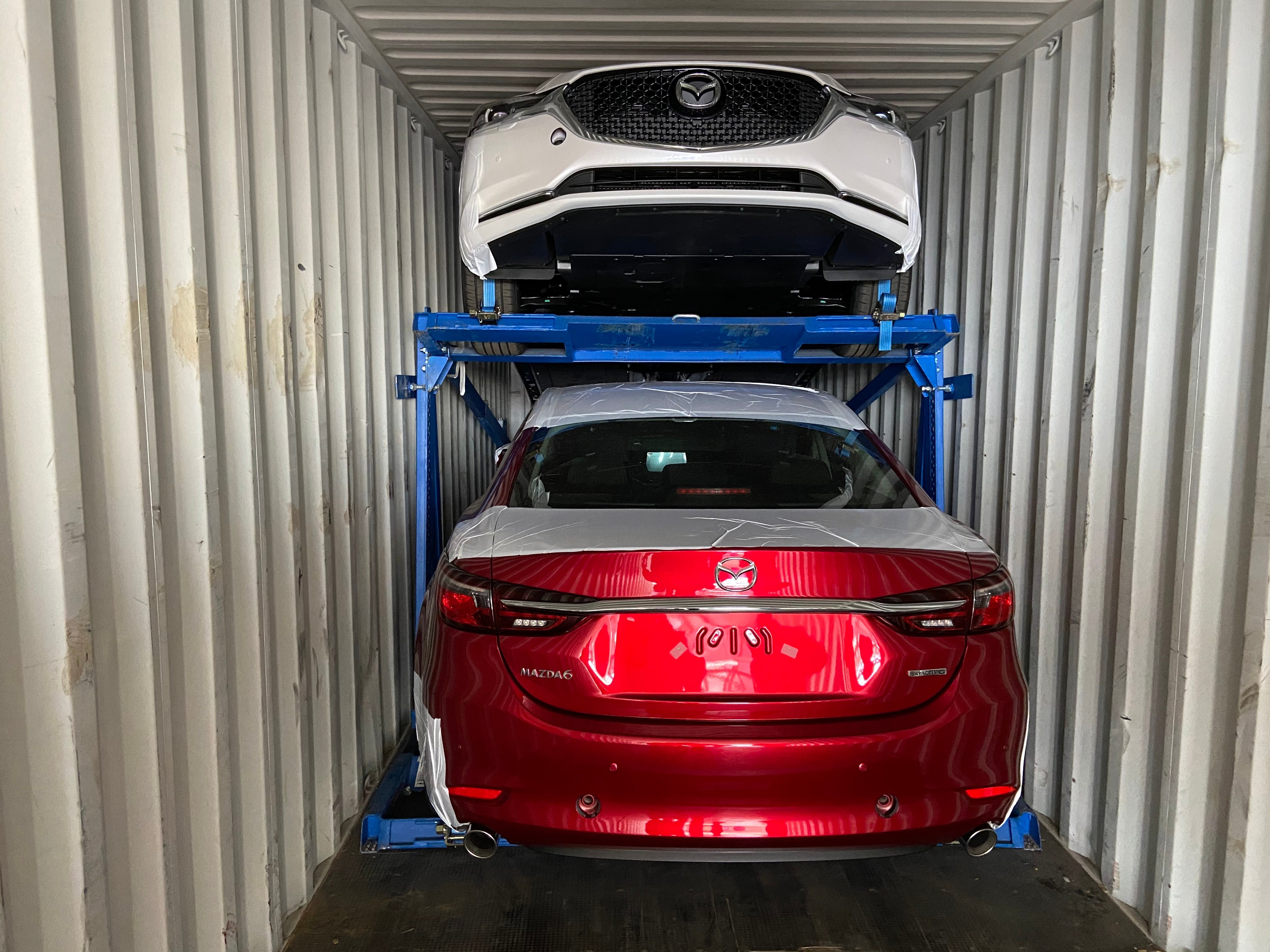 Rear view of a red Mazda 6 sedan and front view of a white Mazda SUV stacked vertically inside a shipping container using a blue Trans-Rak R-RAK steel racking system.