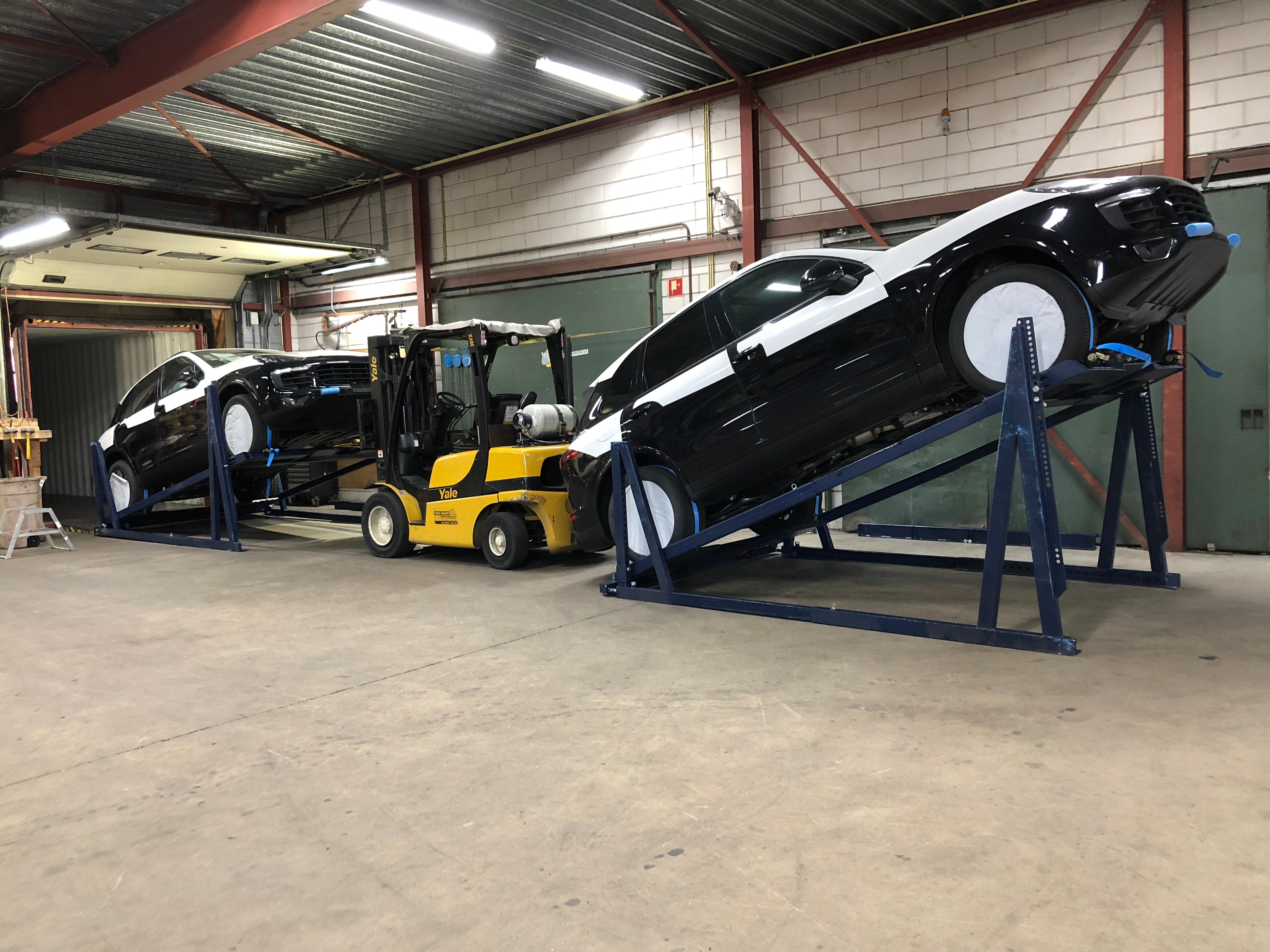 A yellow Yale forklift positioning two black SUVs secured on blue R-RAK automotive racking systems at an angle for container loading.