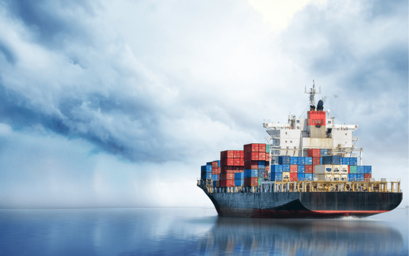 A modern cargo ship laden with colorful containers floats on a calm sea, under a sky where storm clouds loom, symbolizing the critical need for advanced technology in firefighting to protect such vessels on their voyages.